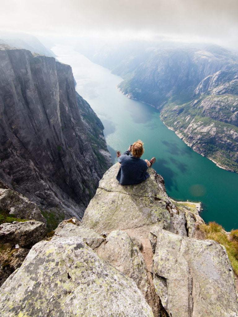 Hiker sitting on Kjeragnasen over Lyseford in Norway taking pictures with smartphone